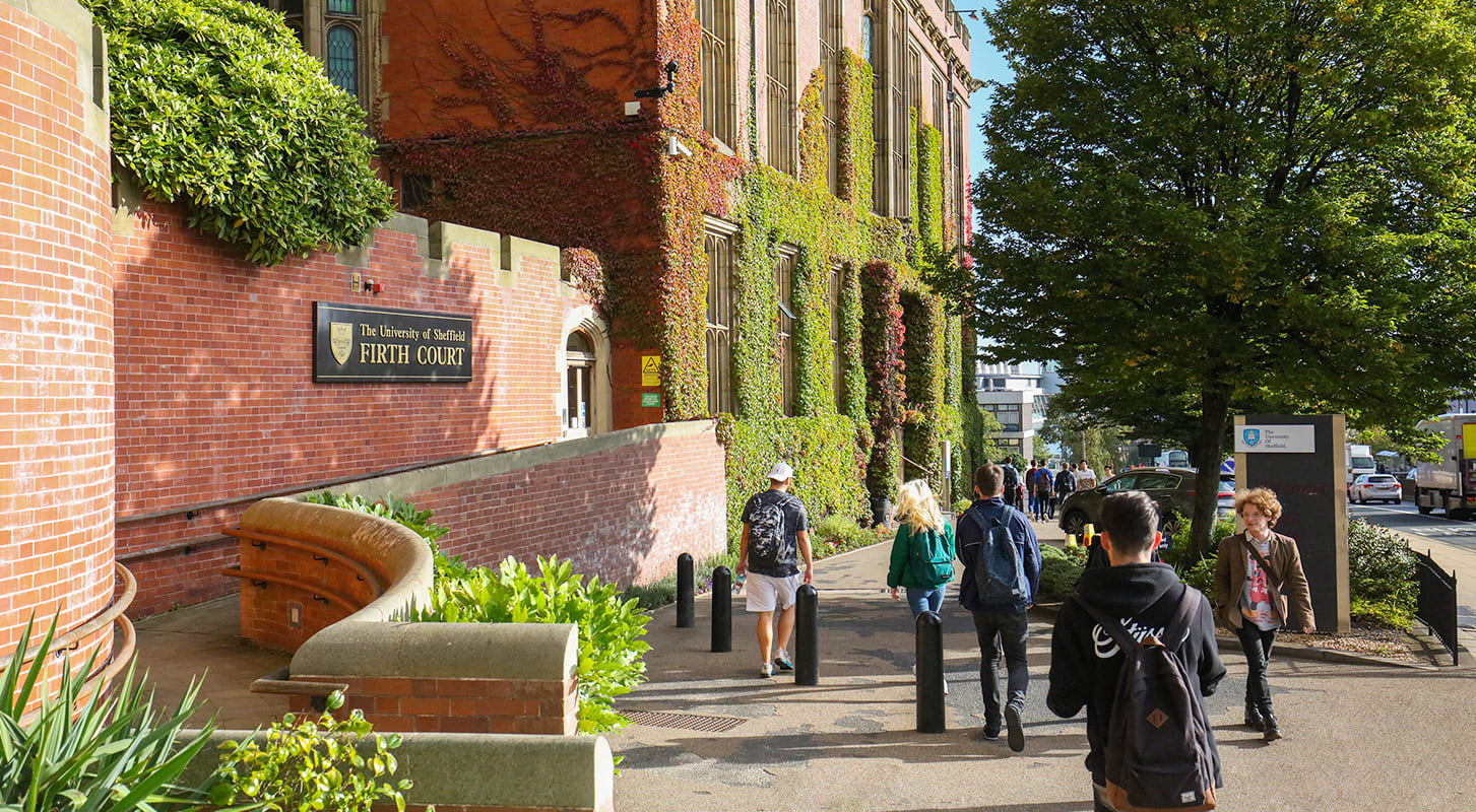 Students with backpacks walking on a sidewalk near a brick building, heading towards the university campus.